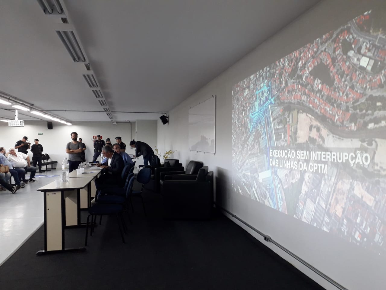 Imagem de uma sala de aula do Centro Universitário Anhanguera com pessoas reunidas ao fundo para a apresentação da audiência. À direita um telão com a projeção de slides
